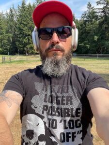 A recent photograph of the author, standing in a field with a beard, wearing sunglasses, a cap, large headphones and a shirt by dashare.zone reading 'IT IS NO LONGER POSSIBLE TO LOG OFF'
