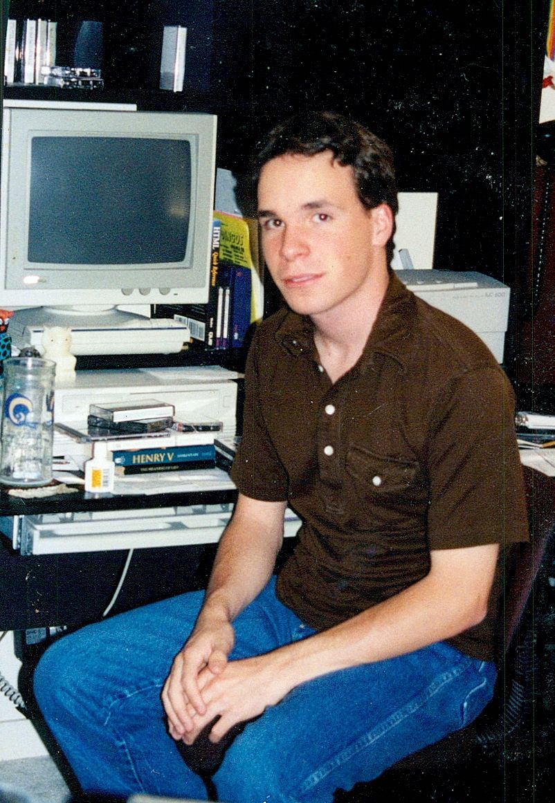 Photograph of the author circa 1998. An 'awkward' teenager in front of a computer desk stacked with books.